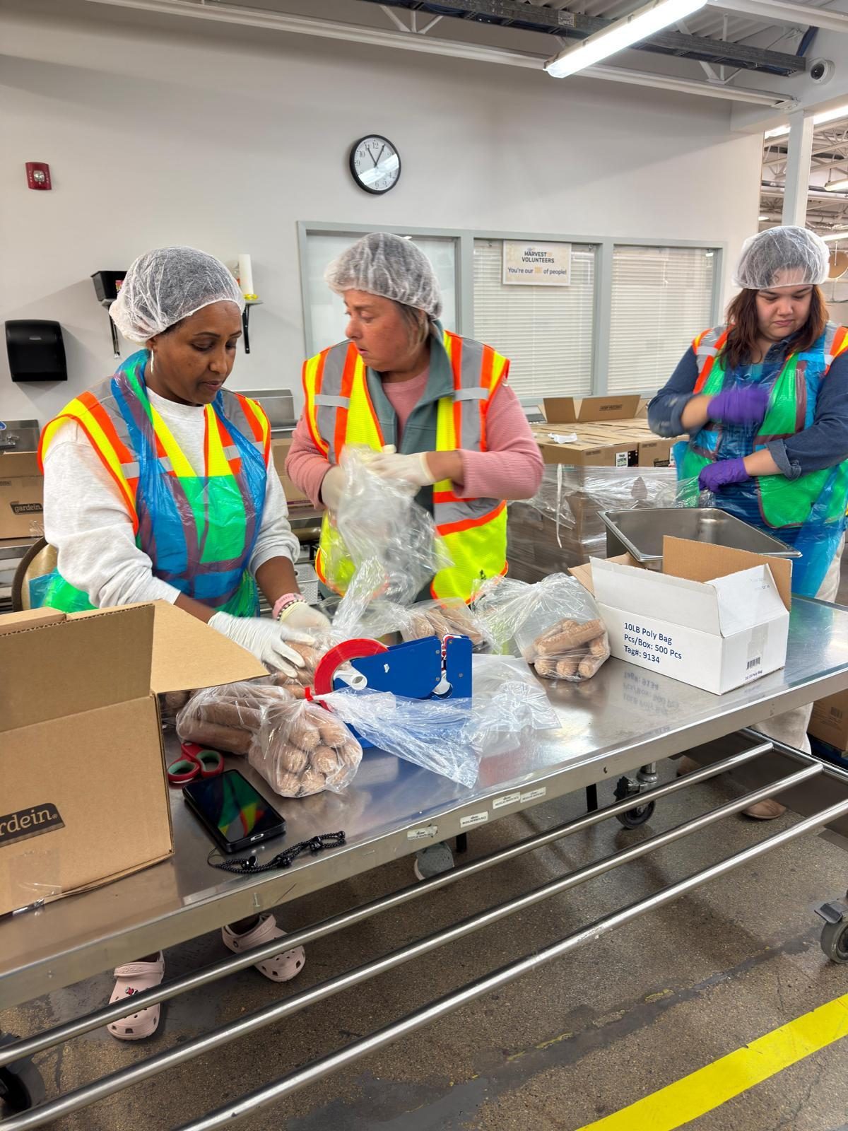 Three participants in hairnets, gloves and safety vests bagging bread together at a warehouse work placement